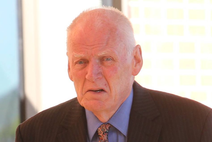 An elderly looking man with thin white hair in a black suit looks at the camera as he walks past.