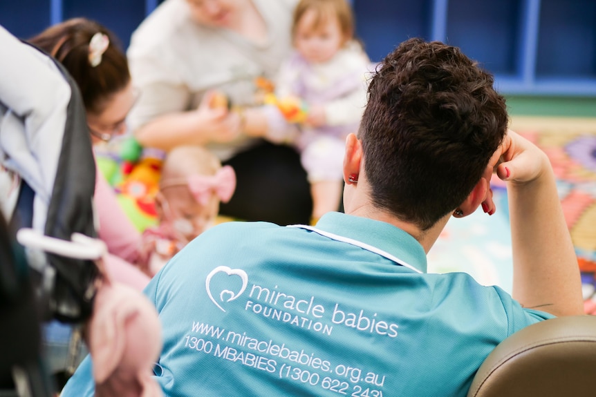 A woman's back of shirt which says Miracle Babies Foundation, with parents in the background.