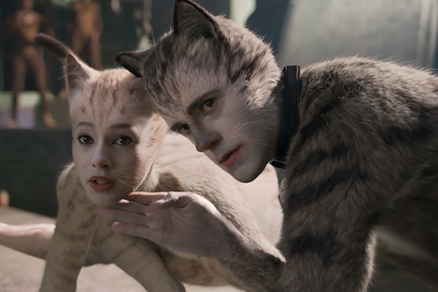 A blue tabby CGI cat with man's face crouches on fours and holds hand under chin of cream coloured CGI cat with woman's face.