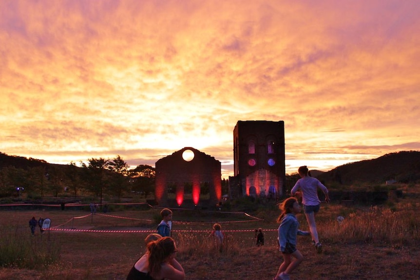 Children running in front of industrial buildings lit up with coloured lights against a dramatic sunset