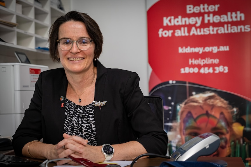 A brunette woman with glasses sitting at a desk smiling next to a Kidney Health Australia Banner 