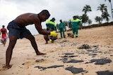 A man is seen bent over with swimming shorts on and yellow gloves on his hands, which he is using to pick up oil clumps on beach