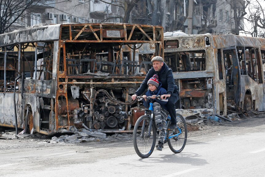 A man and a child ride a bicycle past burnt out buses