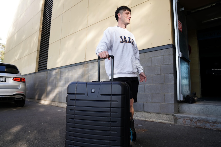A man with brown hair holding a black suitcase, wearing a white jumper with the word Jazz written on it