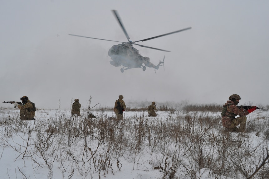 Armed soldiers in the snow as a helicopter hovers closely above.