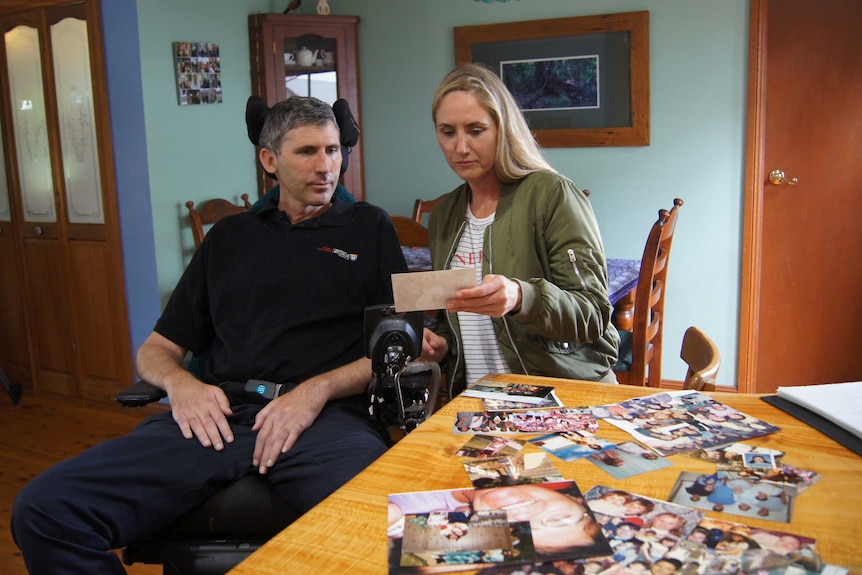 A man in a wheelchair and a woman on a chair look at photographs sprawled out over a counter