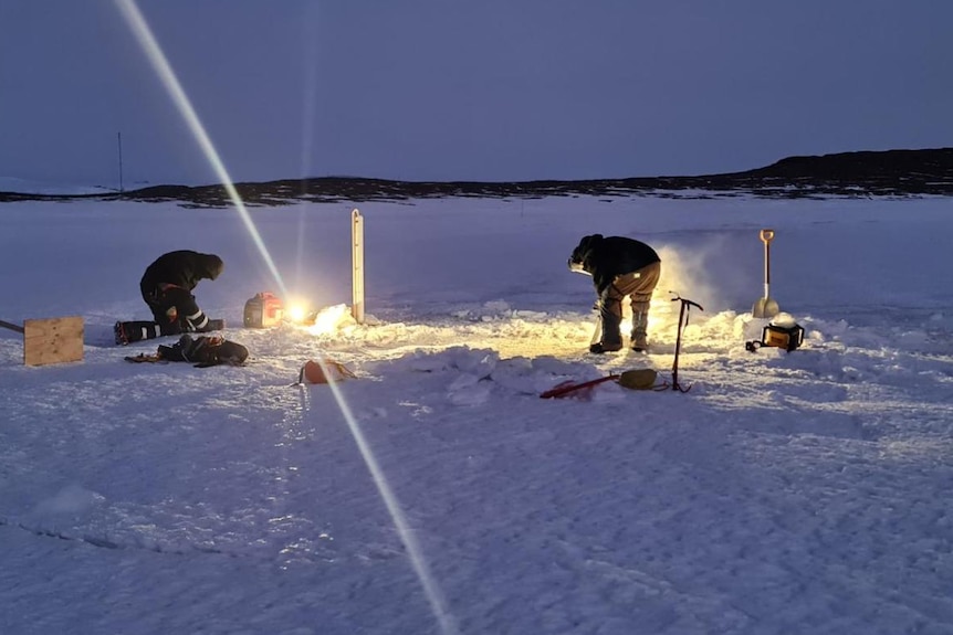 Two people cut a hole in the ice by torchlight at Mawson station, Antarctica, June 2023 