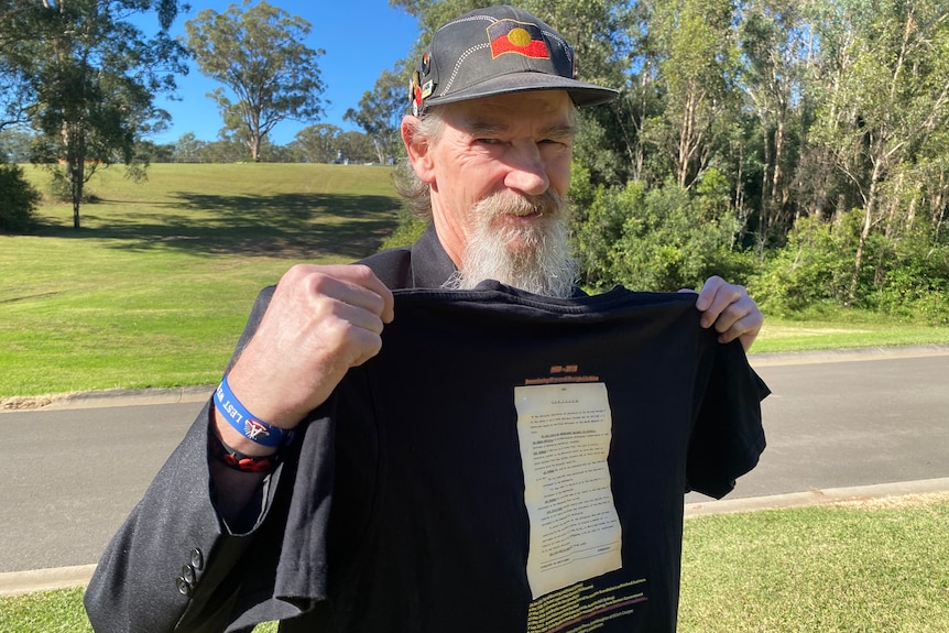 Michael is wearing a black blazer and smiling at the camera. He is holding up a black t-shirt with the petition printed on it