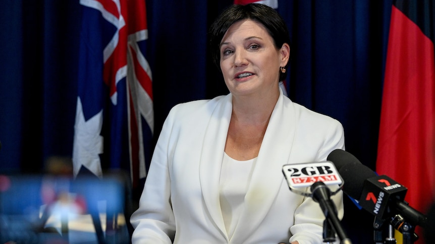 Jodi McKay, wearing a white jacket, stands behind microphones.