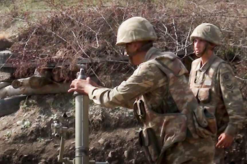 A solider loads a round into a mortar cannon that is pointing upwards while another soldier looks on.