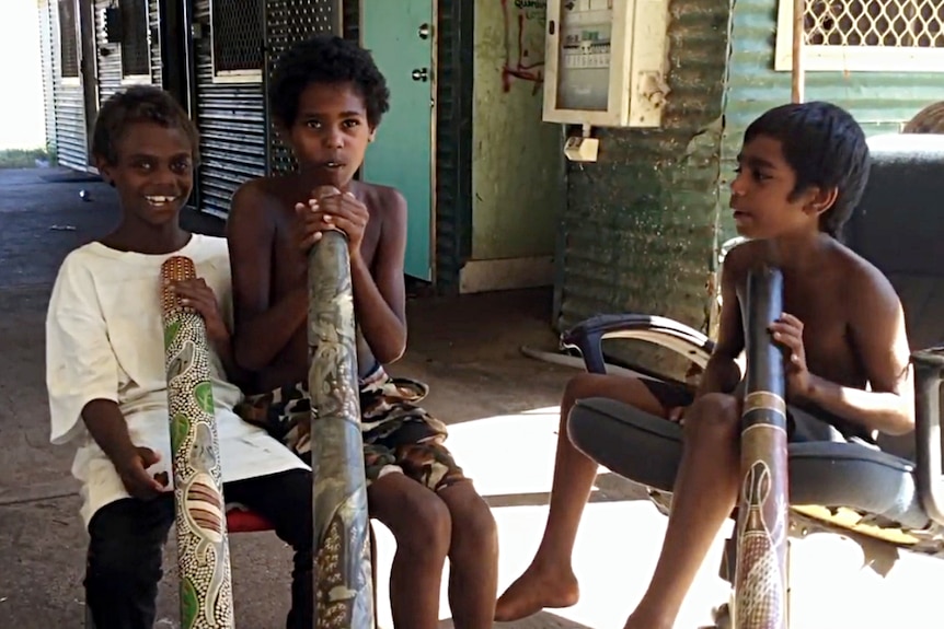 Three children sit on chairs playing didgeridoos.