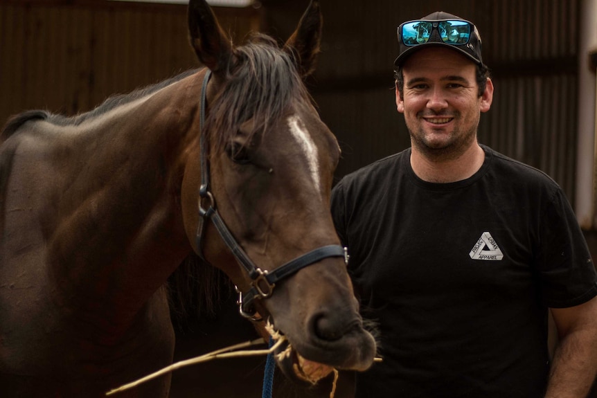 Man in horse stables smiles as he stands behind a horse that is eating hay.