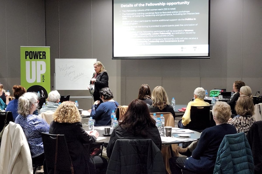 A women leads a talk at a conference, with other women listening, and a powerpoint in the background.