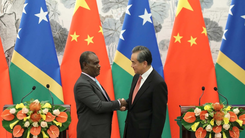 In front of a row of Solomon Islands and Chinese flags, Jeremiah Manele and Wang Yi shake hands in between lecterns.