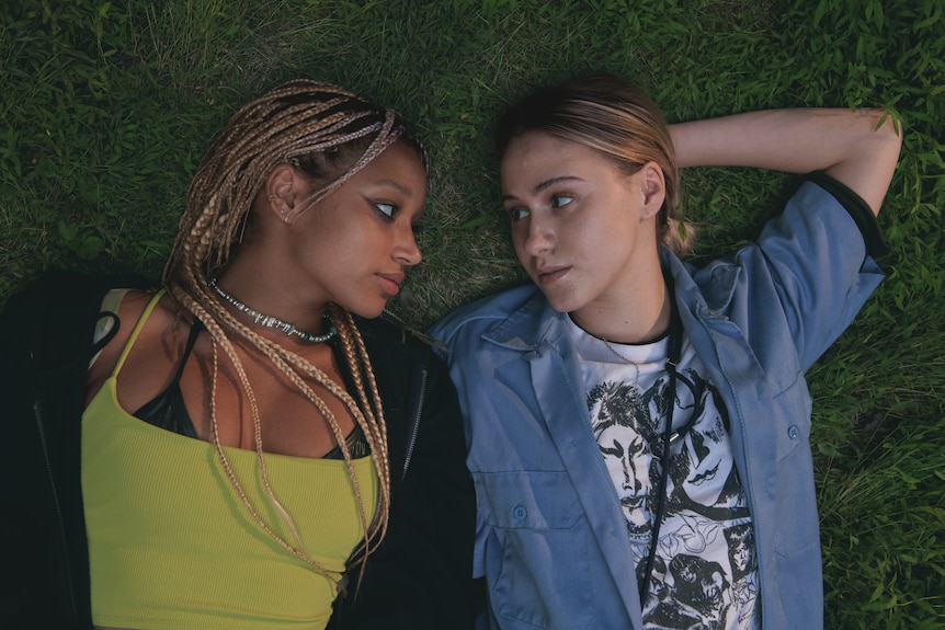 Two women lie on the grass looking at each other.