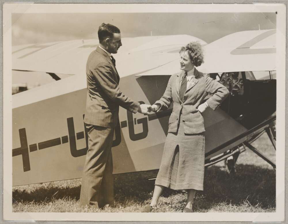 A smiling woman in a skirt suit shakes hands with a man in a suit as they stand next to aeroplane in a field.