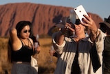 A traveller takes a photo of her champagne with Uluru behind her. There is a large crowd of people in the area.