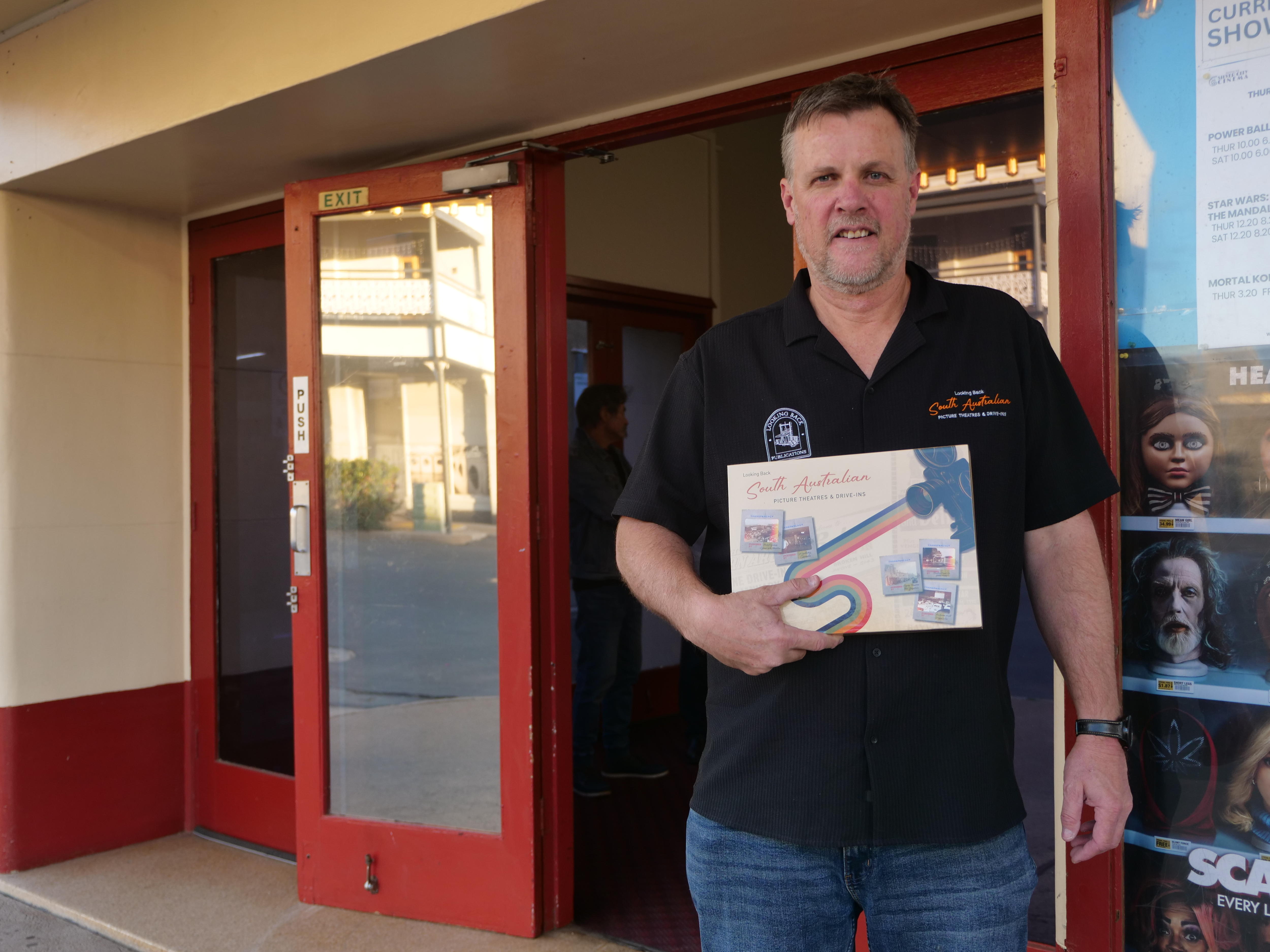 Un hombre se para frente a un cine y sostiene un libro sobre los cines del sur de Australia.
