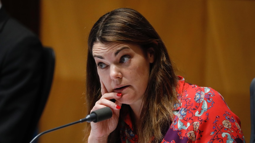 A woman with brown hair wearing a red dress with flowers on it looks disapprovingly with her eyebrows raised