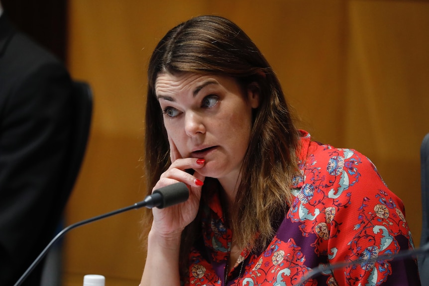 A woman with brown hair wearing a red dress with flowers on it looks disapprovingly with her eyebrows raised