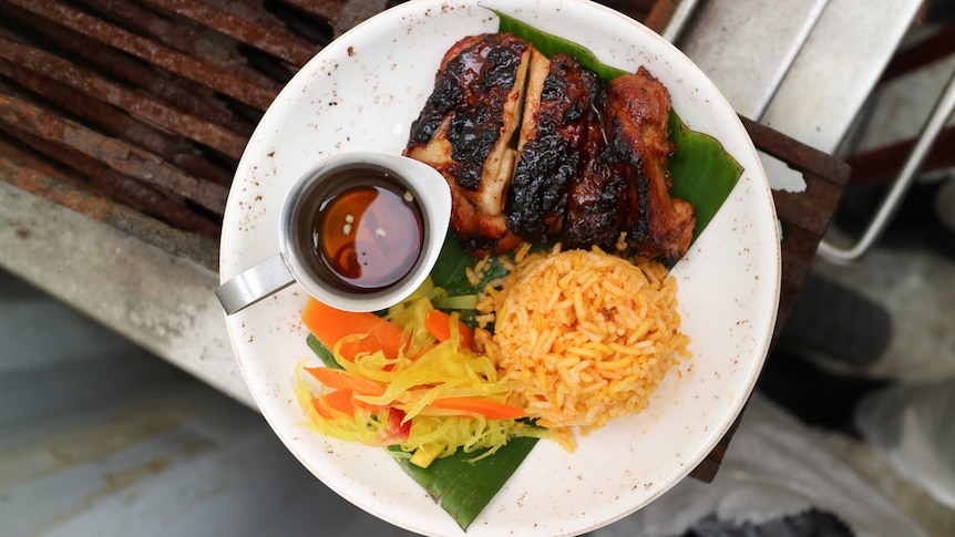 Overhead of a well-barbecued chicken maryland, sauce in metal jug, yellow rice and carrot and cabbage pickles, on a white plate.