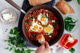 Easy shakshuka with crumbled feta (poached eggs in a spicy tomato sauce) in a pan served on white table with bread.