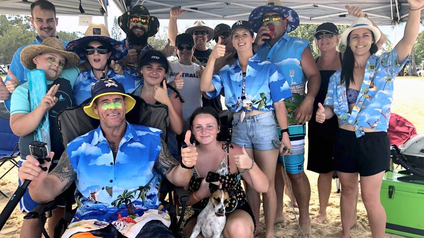 A group of people wearing give the thumbs up as they pose for a photograph under a pop up gazebo on a beach.