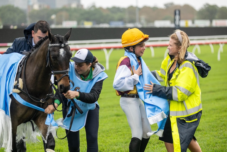 A woman puts a scarf on a girl wearing a jockey outfit next to a horse