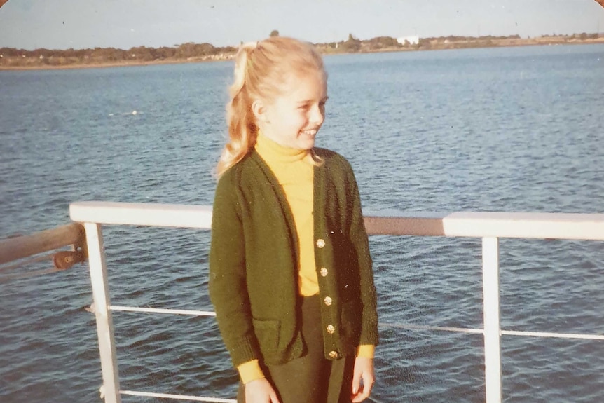 An old photograph of a young blonde girl smiling with her school uniform on a the deck of a boat.