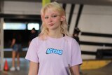 A blonde-haired teenage girl looks coolly at the camera. She's wearing a pink shirt and holding her skateboard.