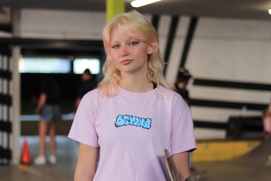 A blonde-haired teenage girl looks coolly at the camera. She's wearing a pink shirt and holding her skateboard.