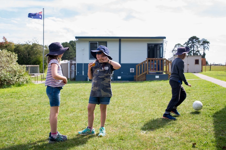 Two girls talk while a boy kicks a ball past them, an Australian flag flying in the background above a small outbuilding.