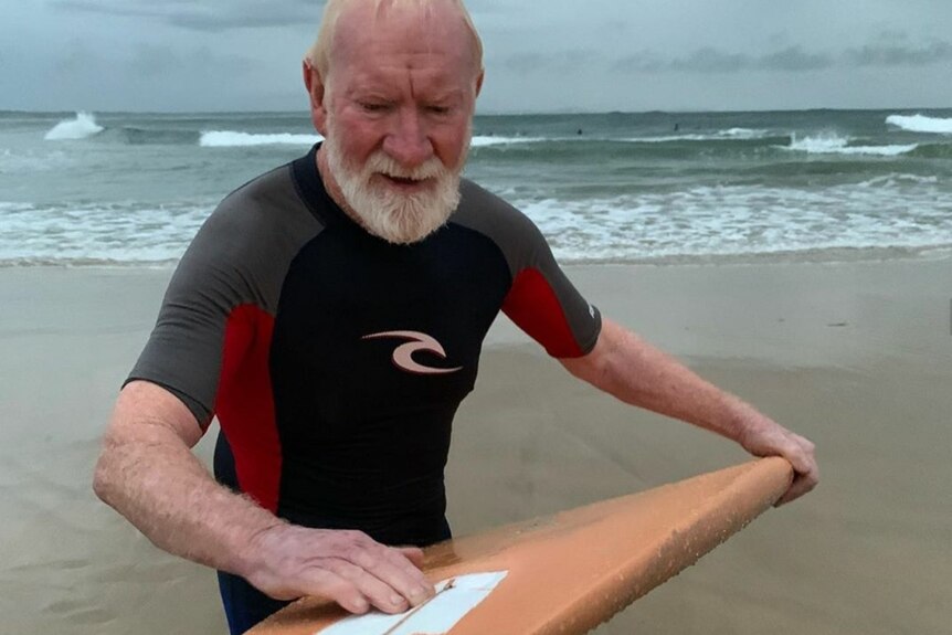 An older man on a beach waxing a surfboard.