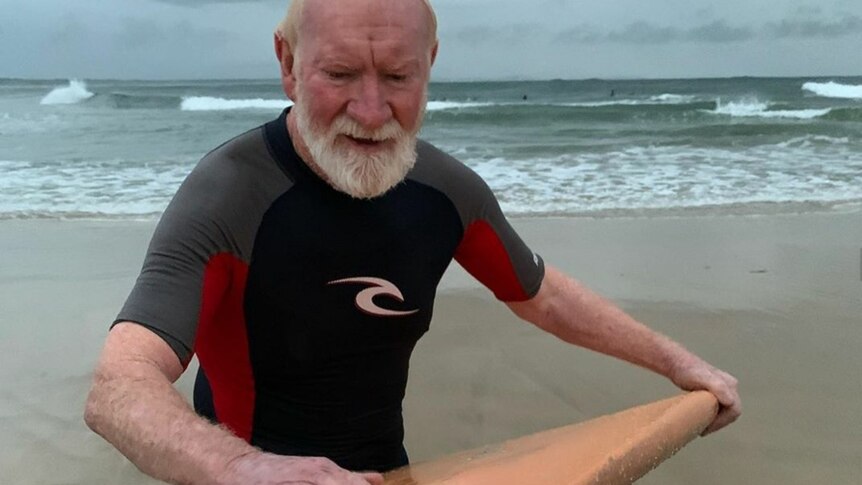 An older man on a beach waxing a surfboard.