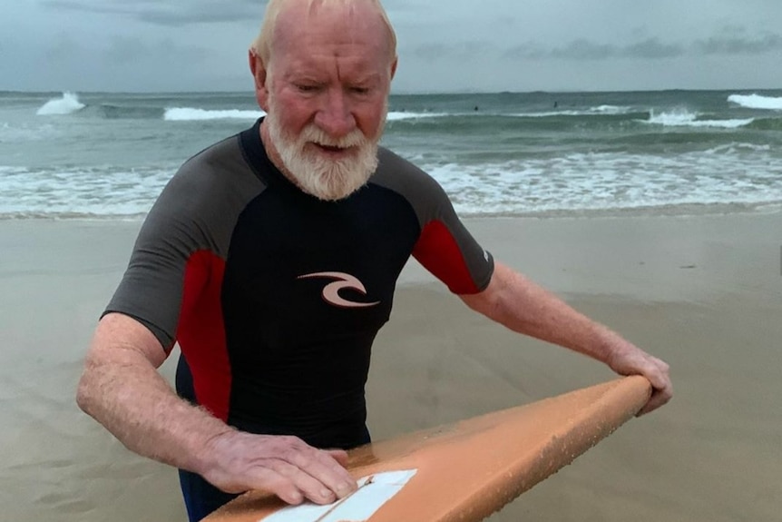 An older man on a beach waxing a surfboard.