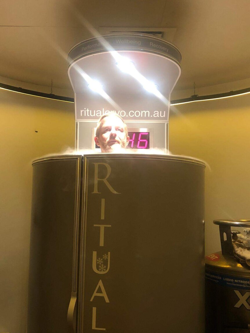 A man's head pokes out from the top of a silver cryotherapy chamber.