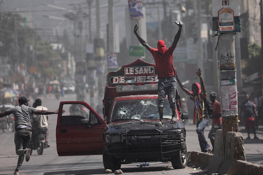 Men hold their hands up on top of a car with bullet holes in the windscreen as men run in the streets