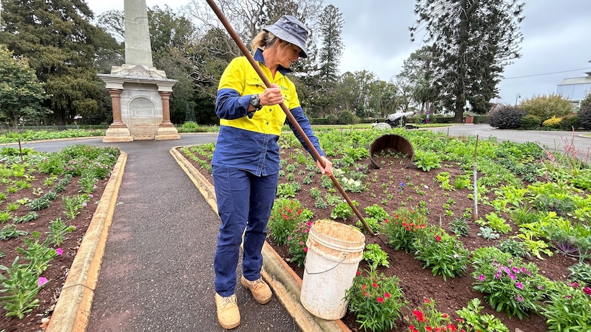 A woman wearing a yellow high-visibility shirt uses a hoe on flowers in a park