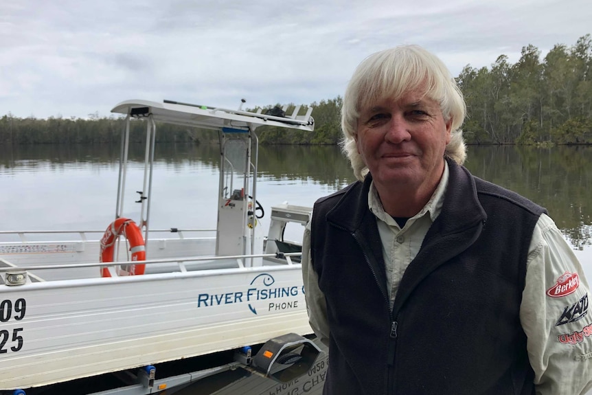 A man in a fishing shirt standing in front of a boat being put into a river.