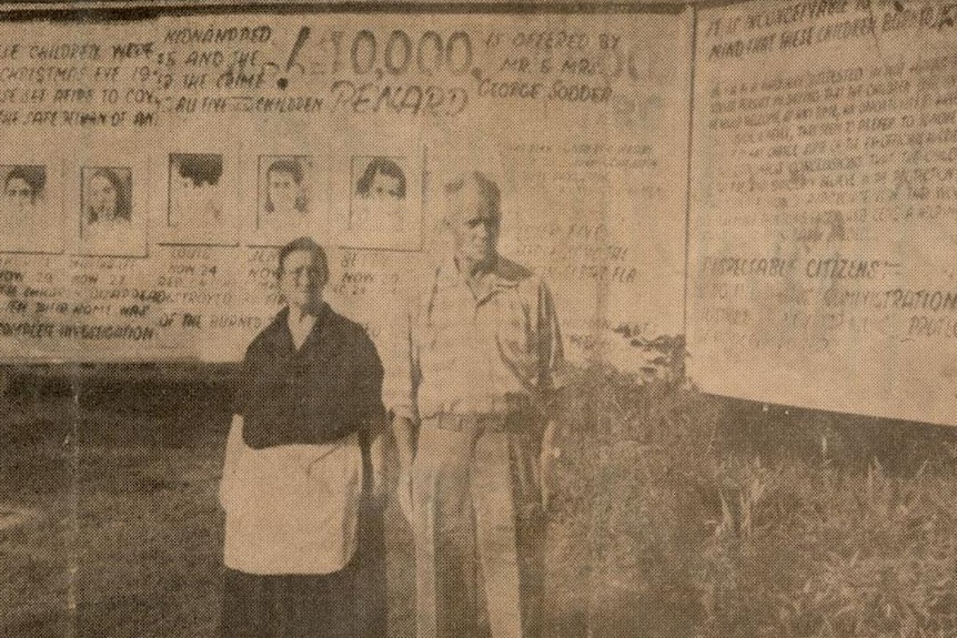 A seipia toned image of a man and a woman standing in front of a billboard displaying five children.
