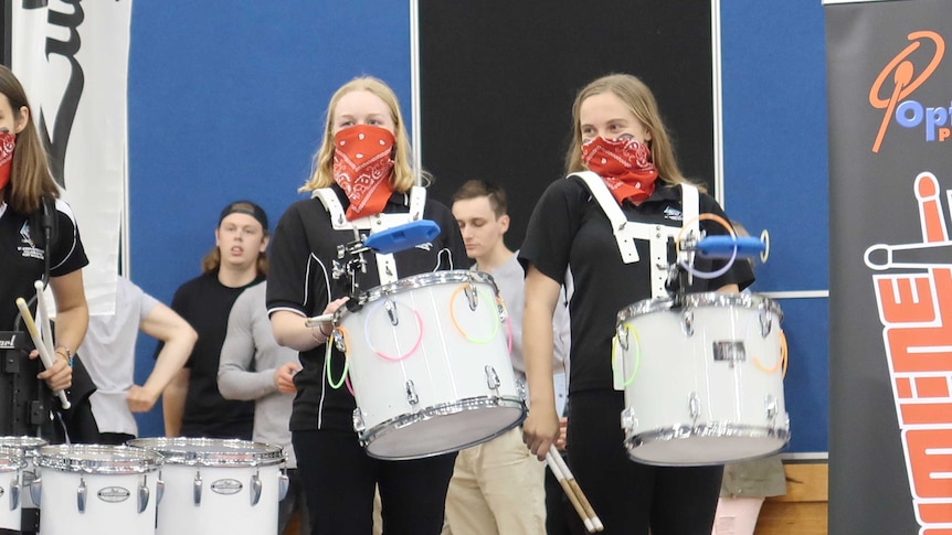 Two girls stand holding white snare drums with red bandanas over their noses and mouths.