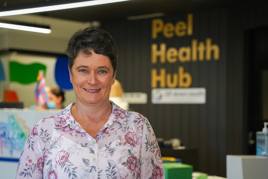 A woman in a flowery blouse stands in front of a sign that says "Peel Health Hub"