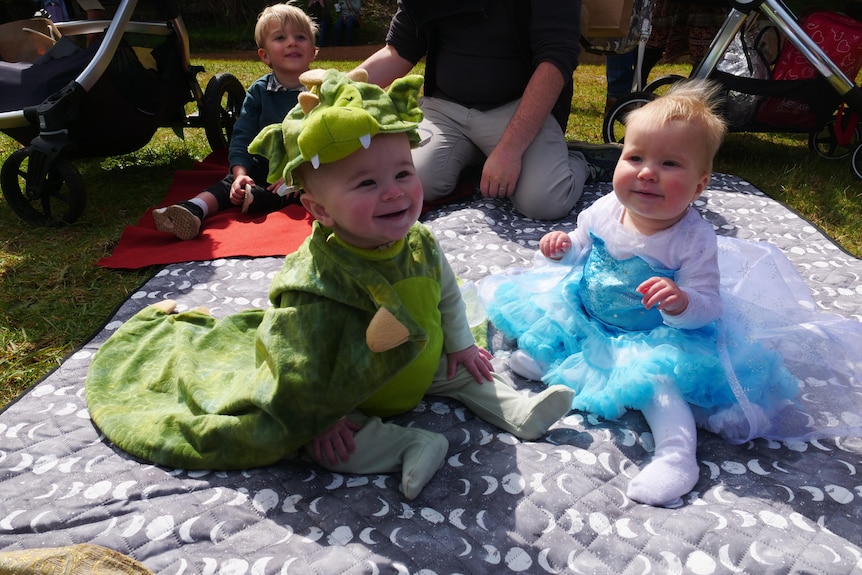 two baby's sit next to each other on a picnic blanket wearing colourful costumes