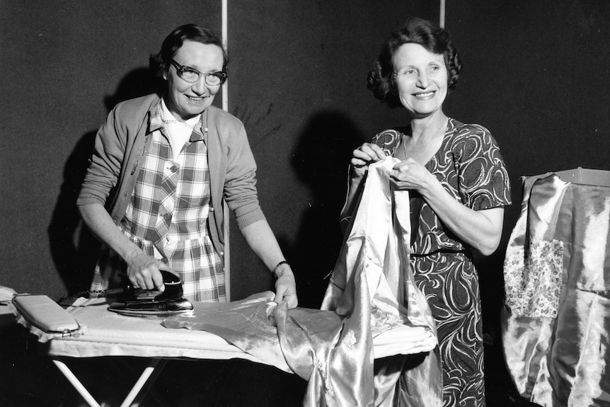 Black and white photo of two women smiling as one irons clothes and the other holds up a shirt with a needle.