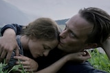A woman with eyes closed and man with serious expression lay in an embrace on grass in the countryside on an overcast day.