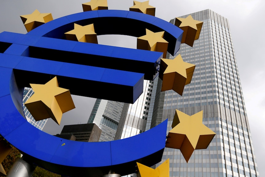A statue of a blue euro symbol surrounded by gold stars, with a tall building behind it.