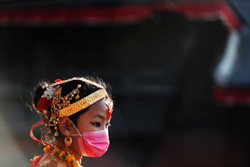 A young girl wearing gold headband and jewellery and a pink mask looks down.