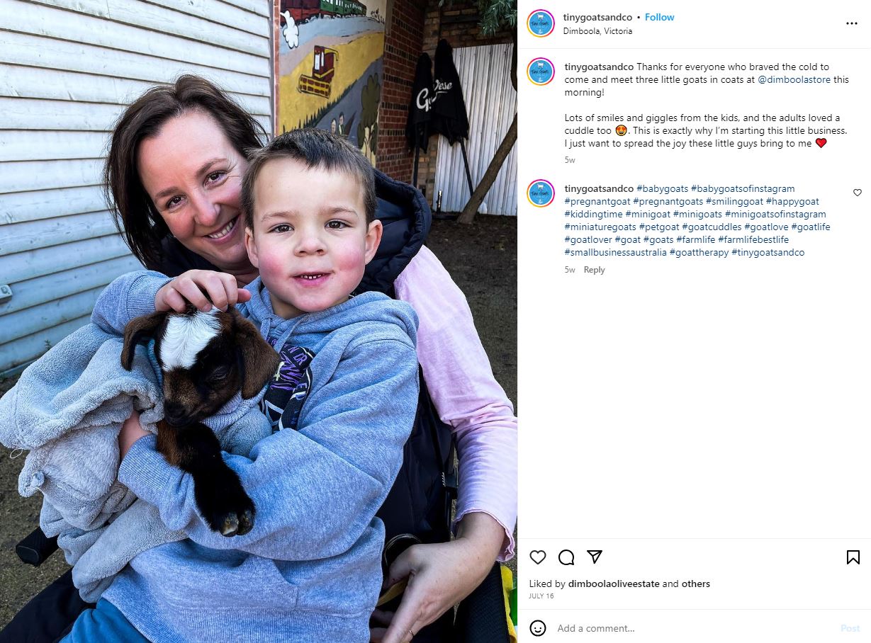 A woman smiles behind a boy holding a little brown and white goat.