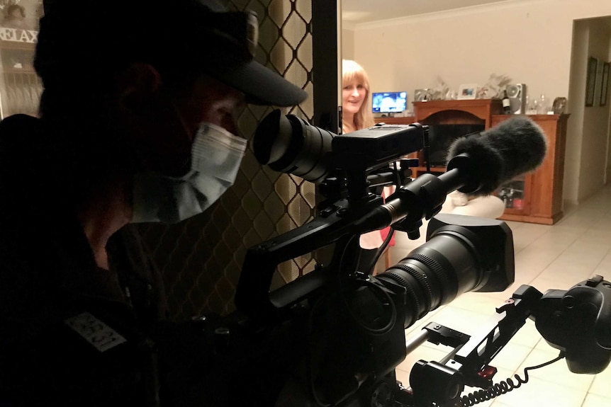 Cameraman wearing mask holding camera filming through doorway to woman inside.