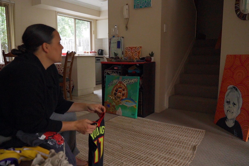 A woman sits in her home looking out to the distance with a vacant expression. She holds her young son's football jersey.